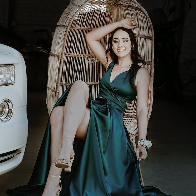 Woman in emerald green gown poses in wicker chair next to a white car.