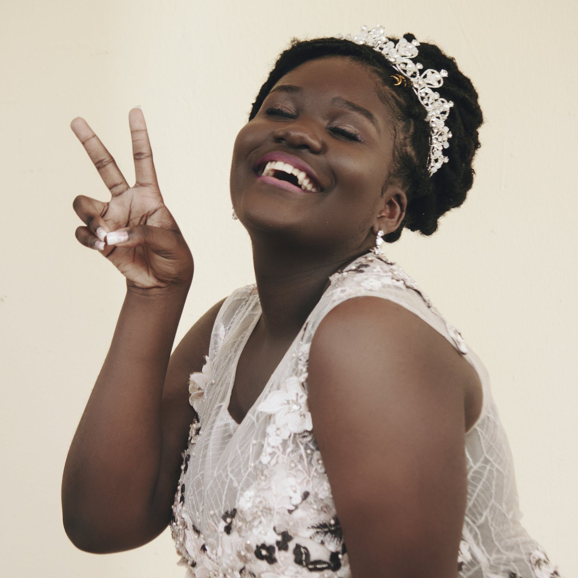 Smiling person making a peace sign, wearing a white dress and tiara, against a beige backdrop.