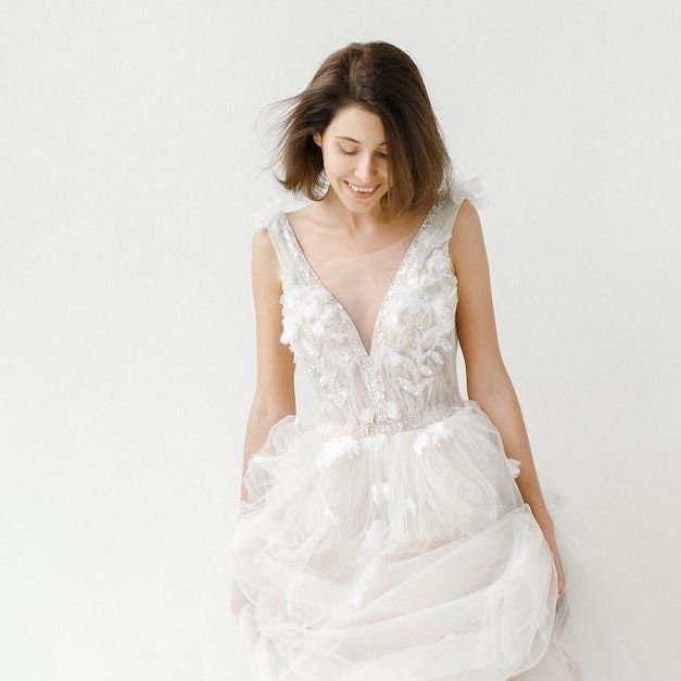 Woman in a white wedding dress smiles, looking down. The dress has a low neckline and tulle skirt. White background.