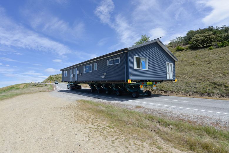A house is being transported on a trailer along a rural road under a blue sky.