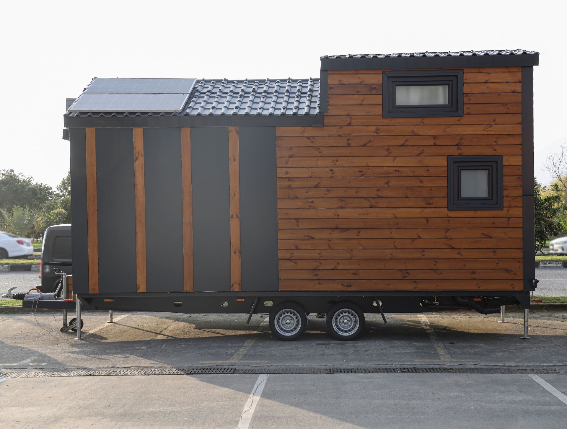 Tiny house on a trailer, black and wood siding, solar panel on the roof, parked in a lot.