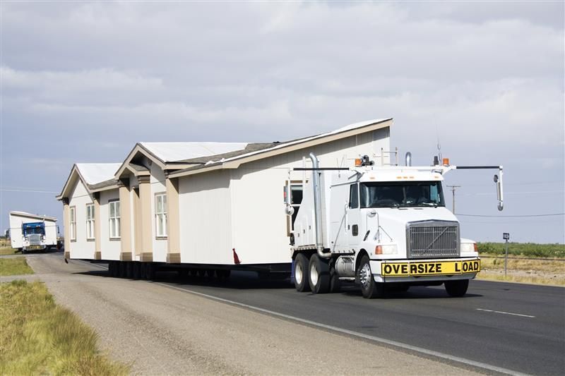 White semi-truck hauling a mobile home on a rural road, under a blue sky.