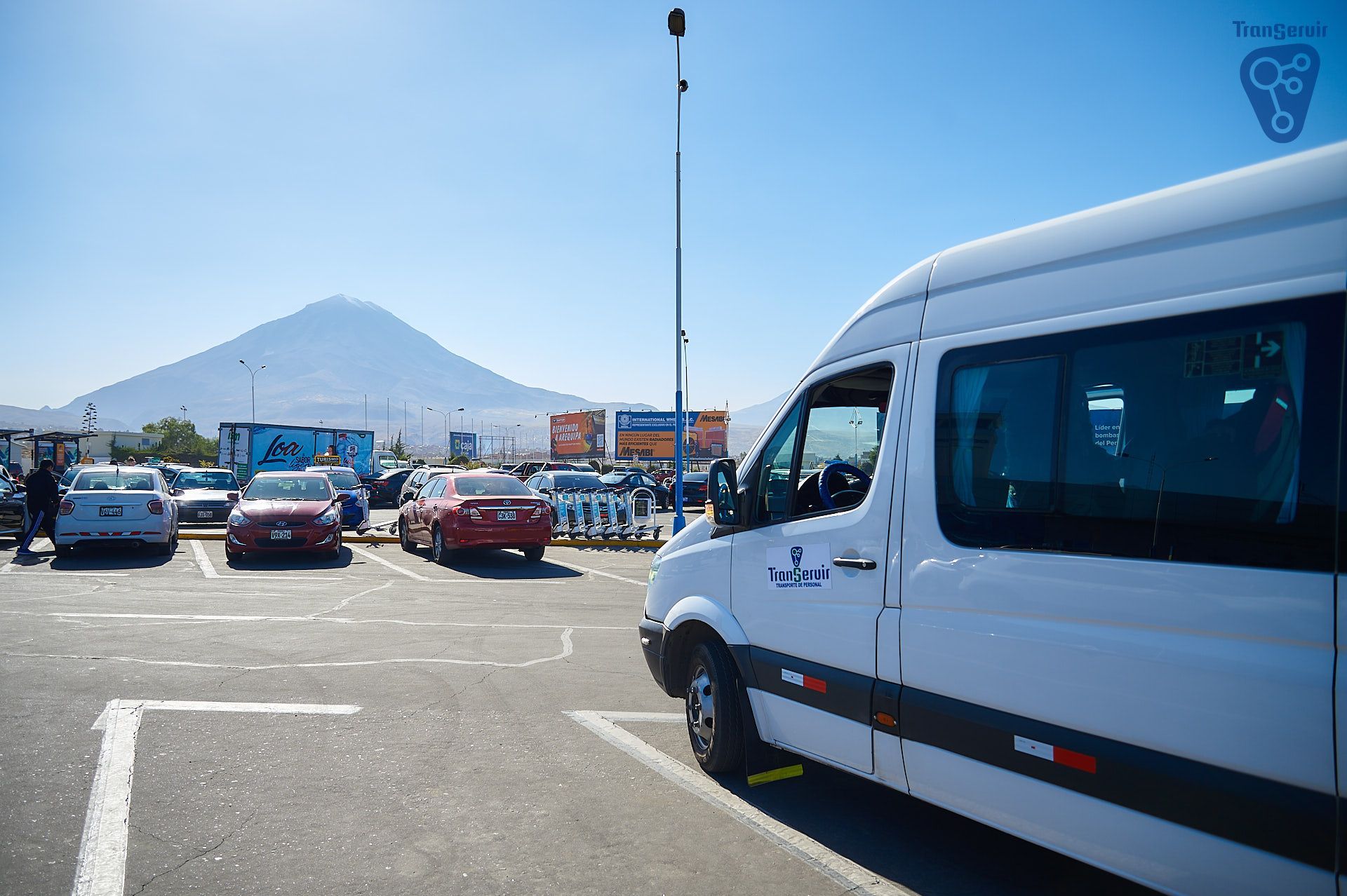 Furgoneta blanca en un estacionamiento con coches, cielo azul y una montaña al fondo.
