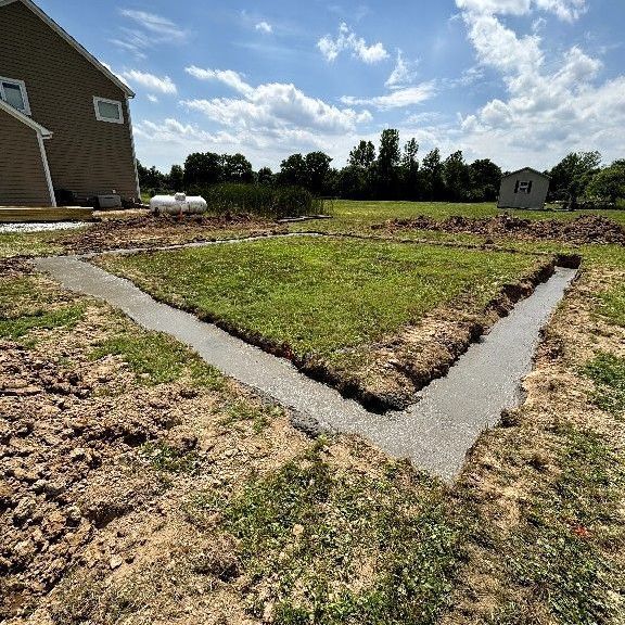 A concrete foundation forms a square on a grassy lawn; a house and shed are in the background.