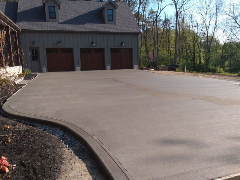Newly poured concrete driveway in front of a three-car garage, with a curved edge and surrounding landscaping.