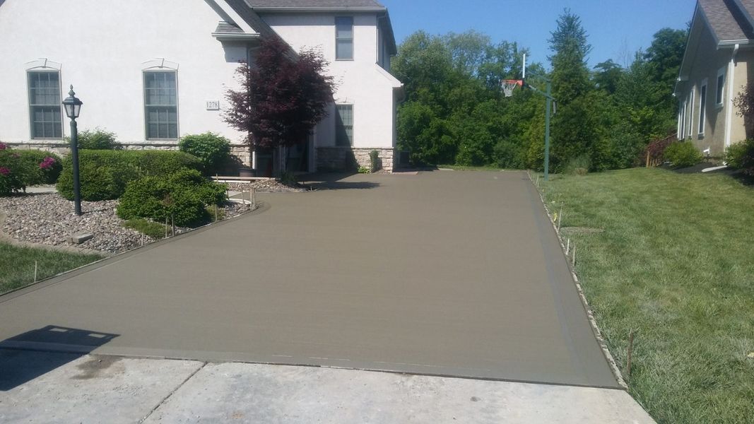 Newly poured concrete driveway in front of a two-story house with green lawn and bushes.