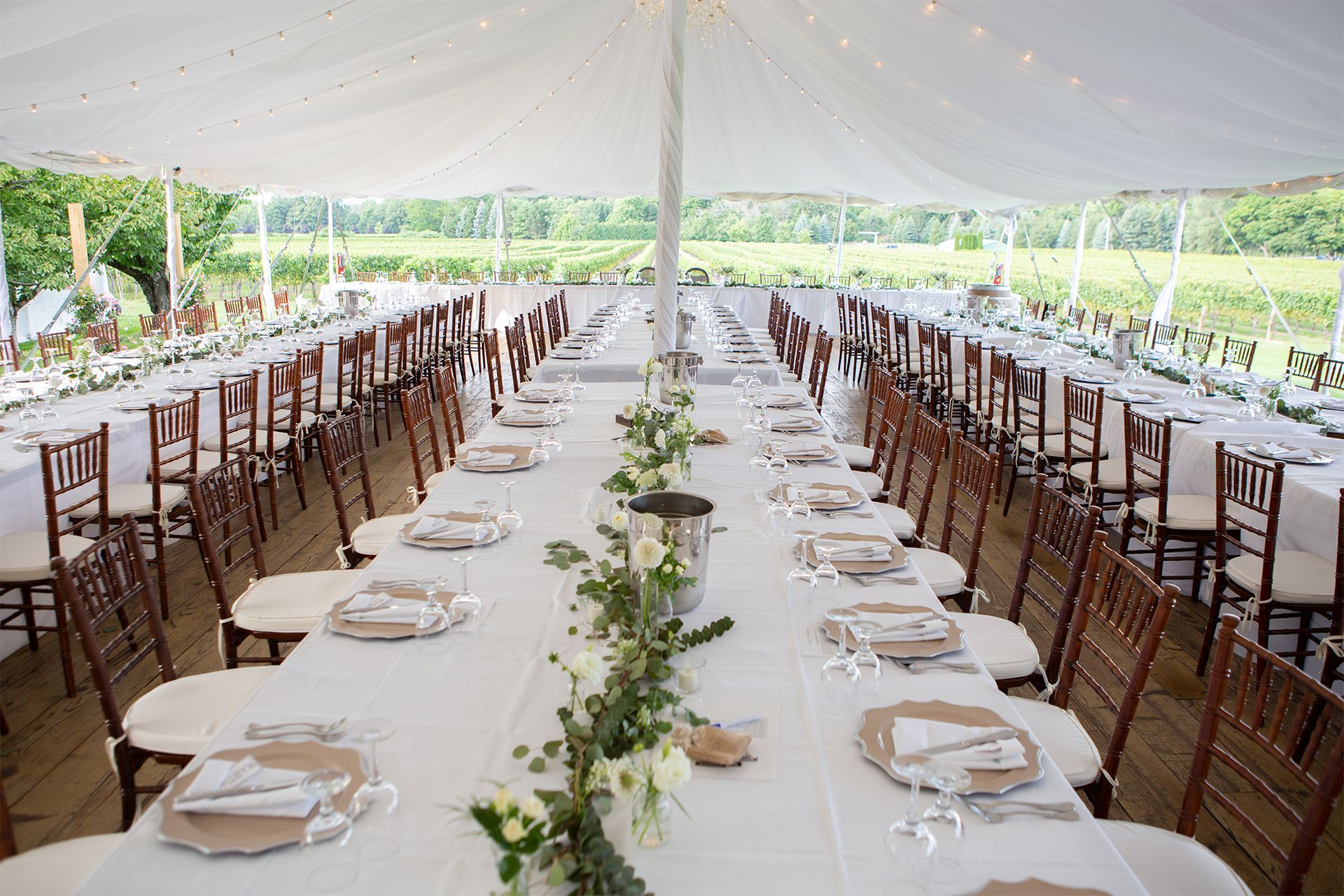 Long banquet table under a white tent, set with plates, napkins, and greenery for an outdoor event. Long banquet table under a white tent, set with plates, napkins, and greenery for an outdoor event.