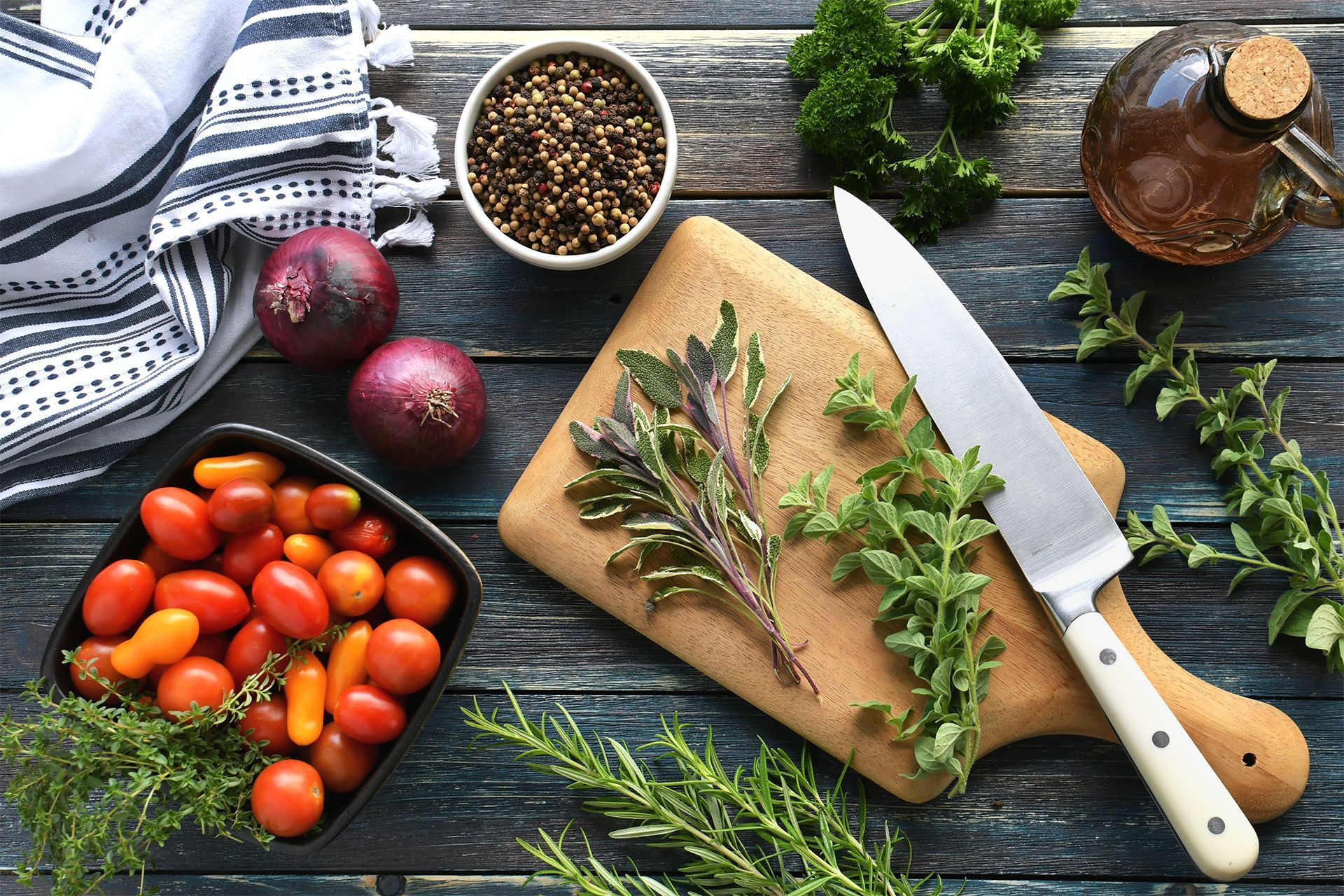 Herbs, tomatoes, onions, knife, and cutting board on a rustic table with fresh ingredients.
