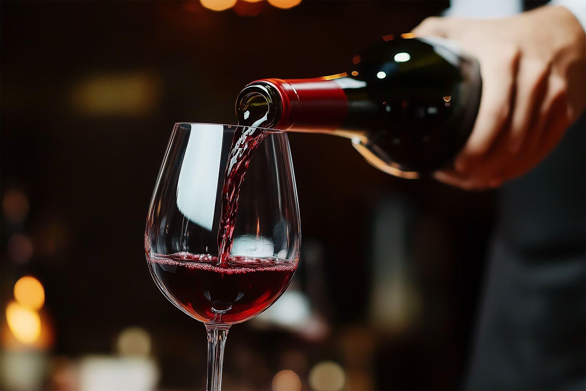 Red wine being poured into a wine glass in a dimly lit setting.