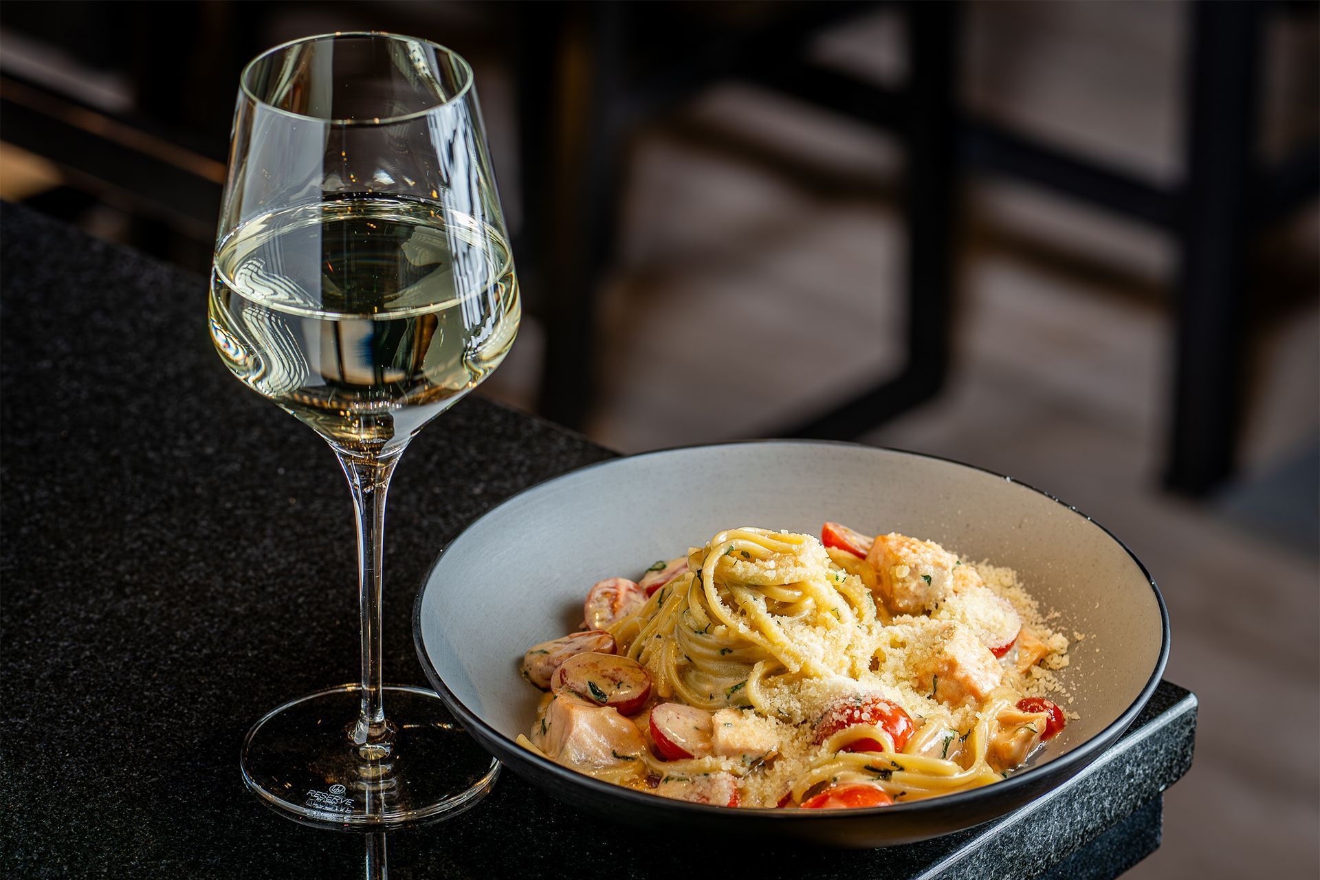 Glass of white wine beside a bowl of creamy pasta on a dark table.