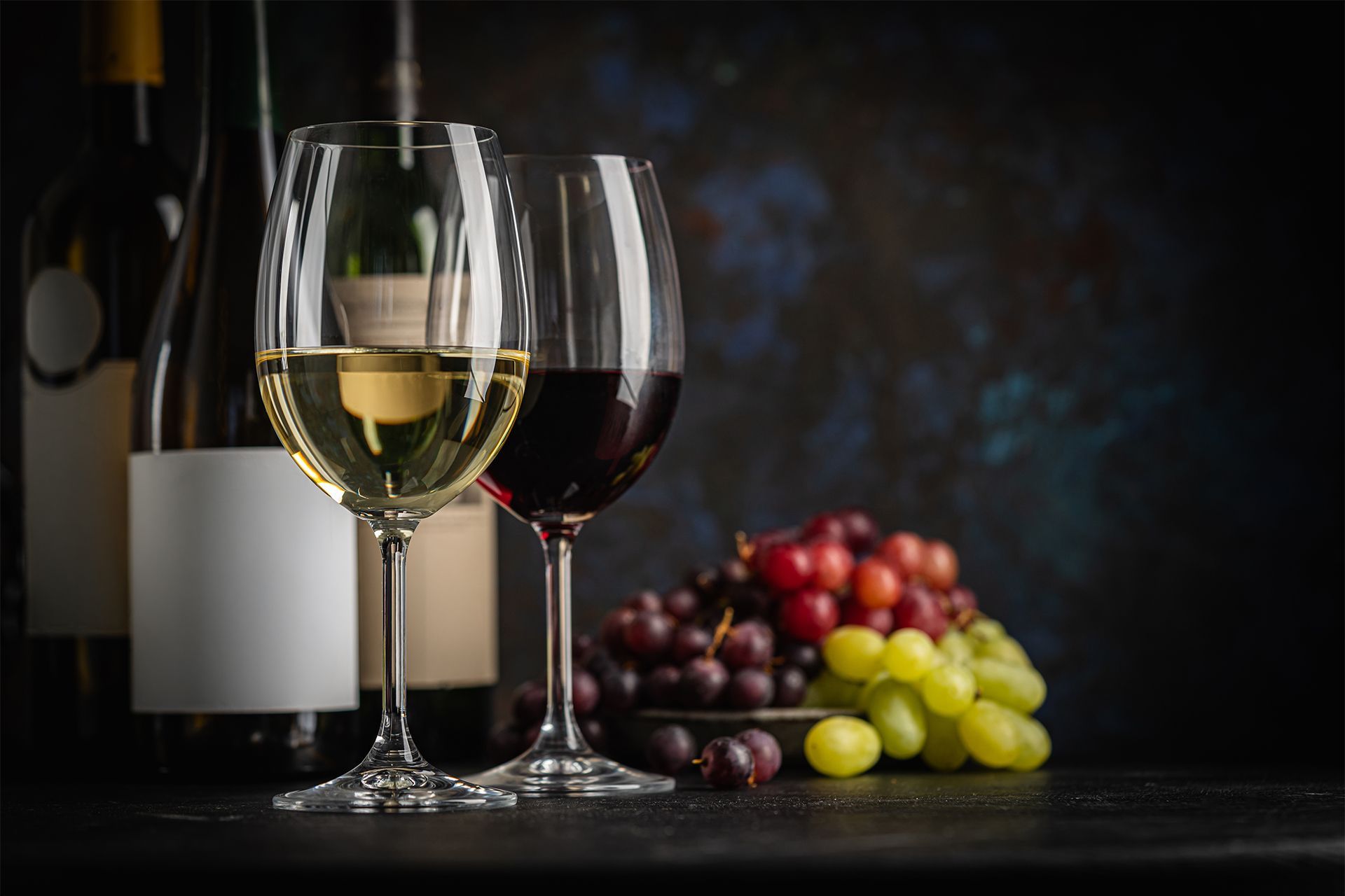 Wine glasses with red and white wine beside bottles and clusters of grapes on a dark table. Wine glasses with red and white wine beside bottles and clusters of grapes on a dark table.