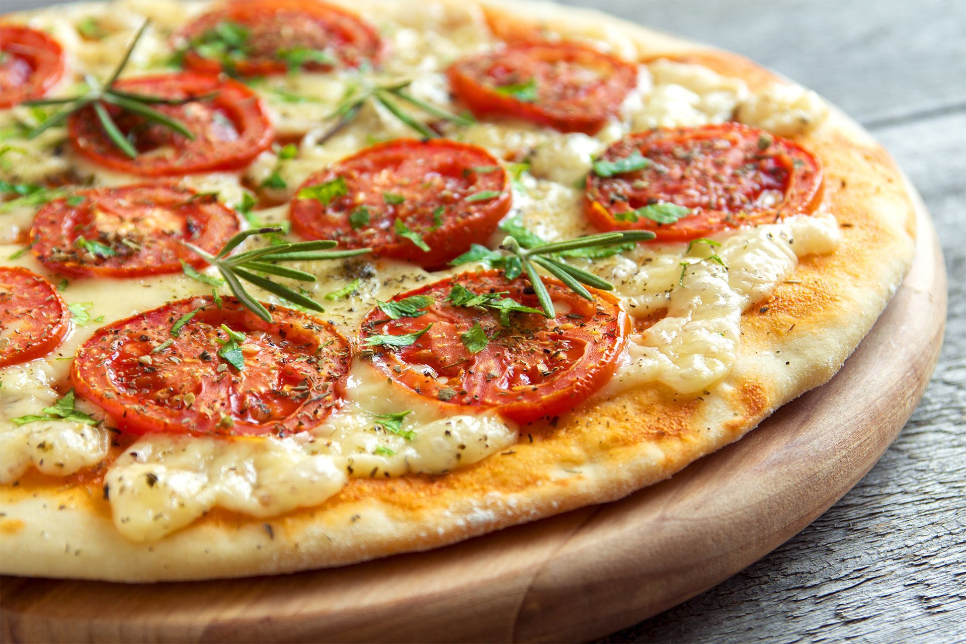 Close-up of a tomato and rosemary pizza on a wooden board, with melted cheese and golden crust.