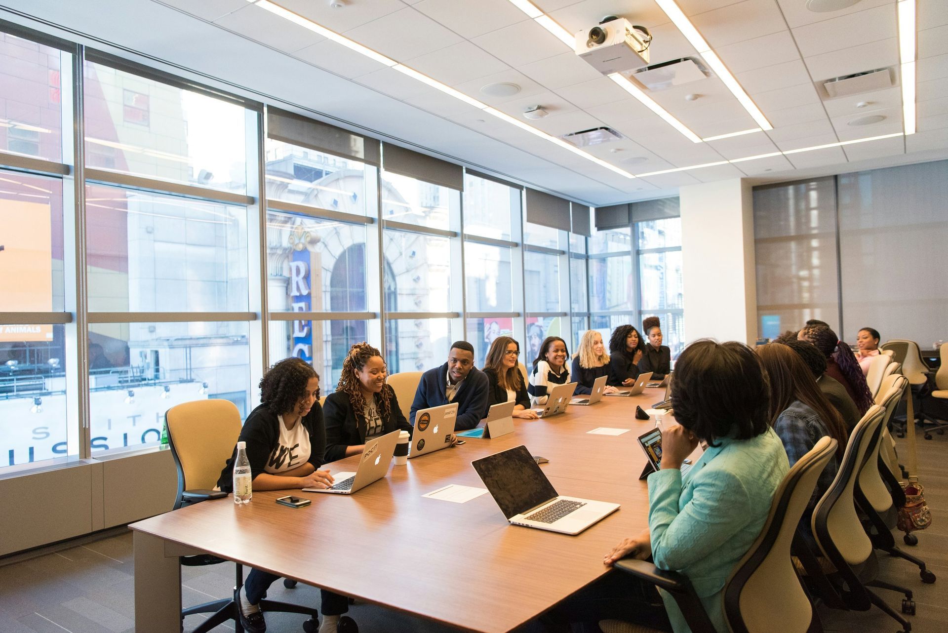 A group of people are sitting at a long table in a conference room with laptops.