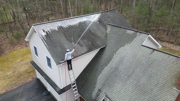 A man is standing on a ladder cleaning the roof of a house.