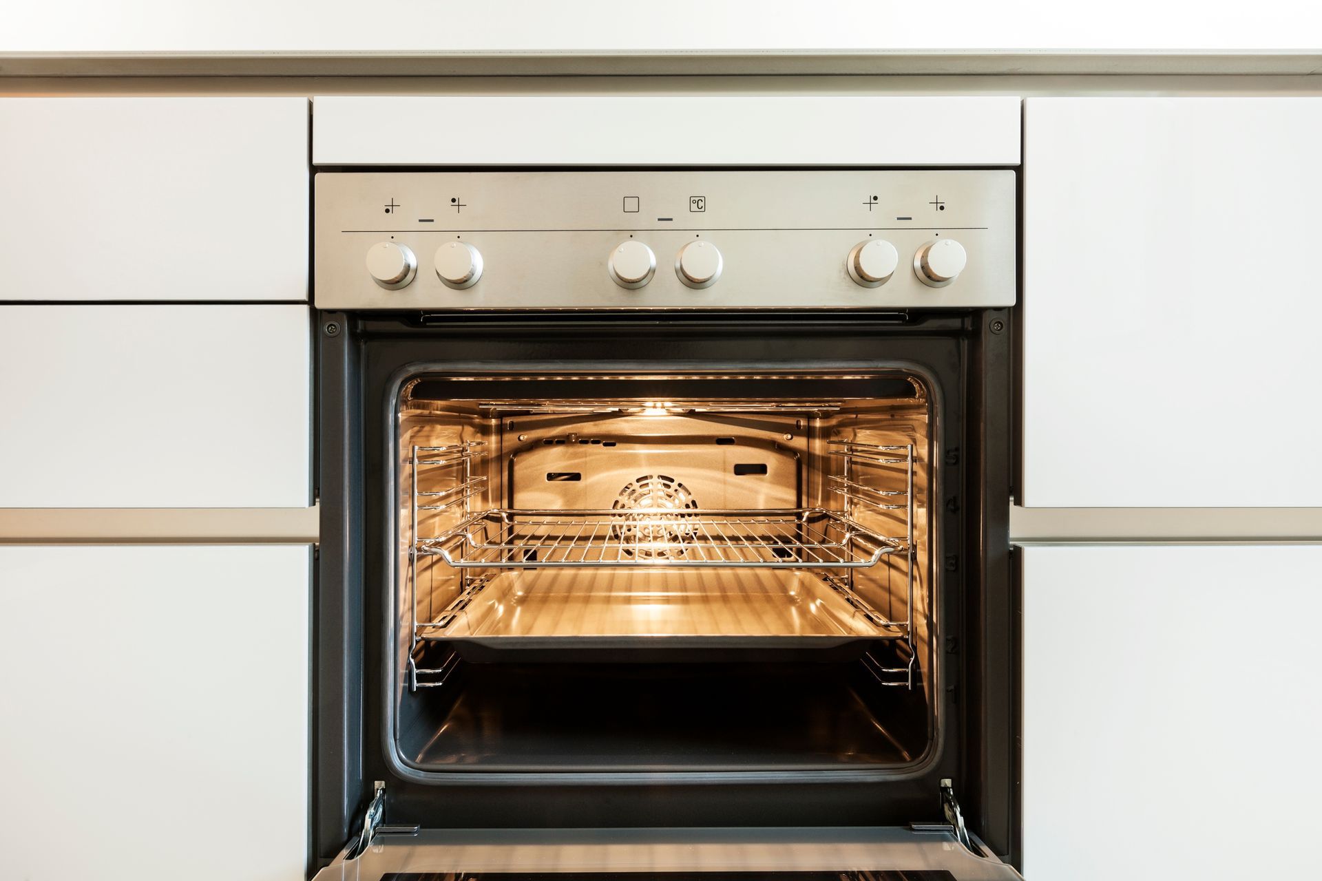Open oven in a white kitchen, interior light on. Silver oven controls.
