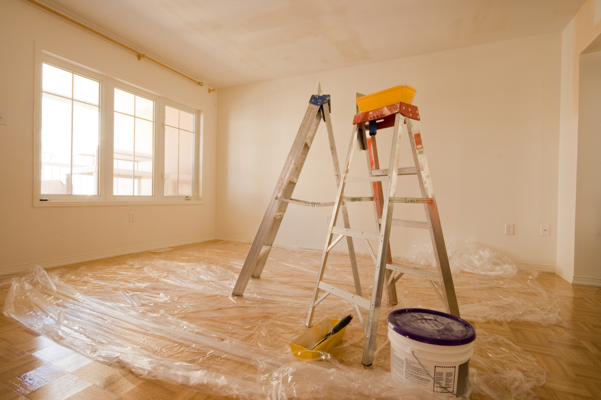 Renovation room with plastic-covered floor, two ladders, and a paint bucket near a window