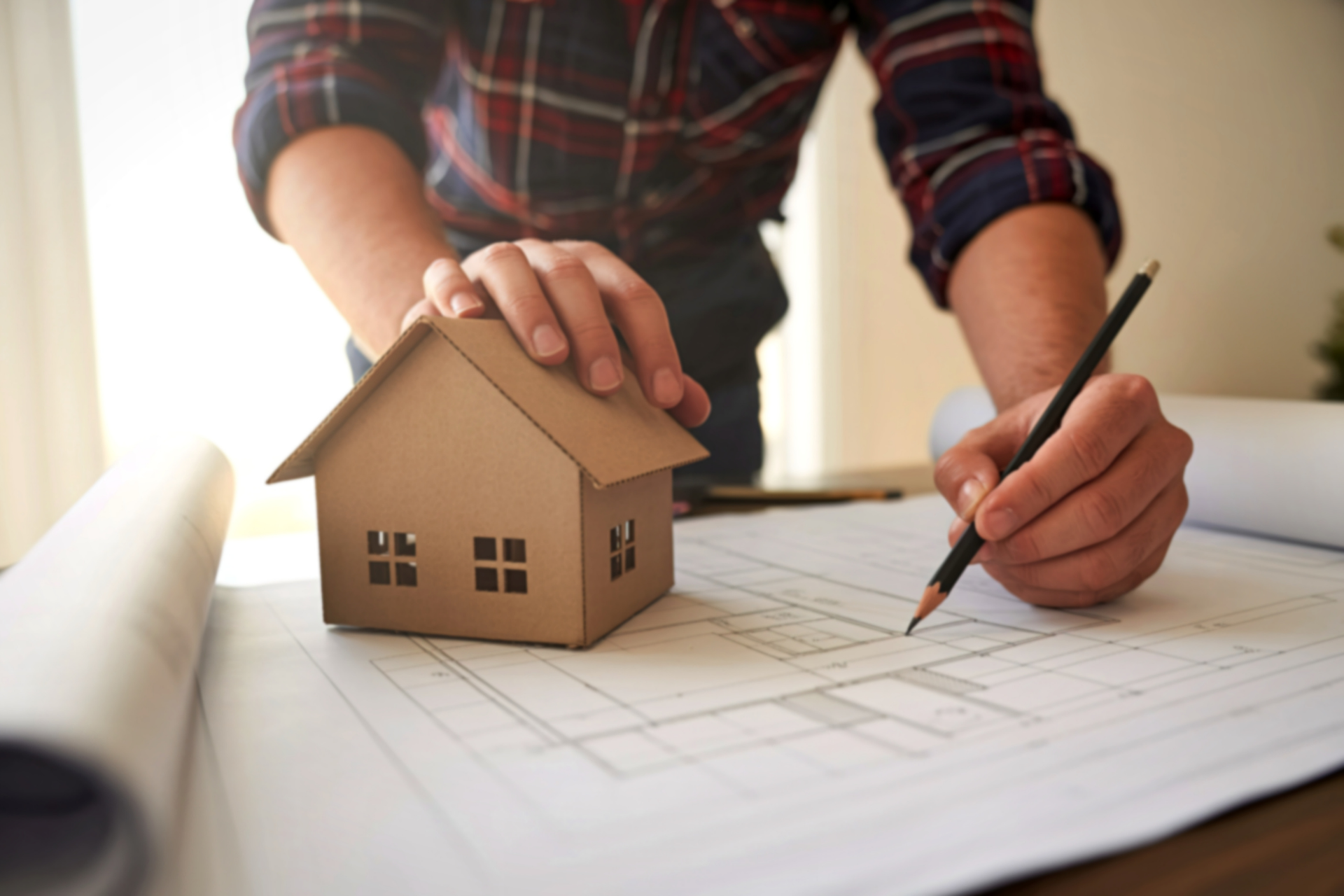 Hands sketching a small house model on a blueprint, with a pencil and ruler on a table