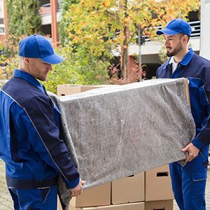 Two Men In Blue Uniforms Carrying A Large Piece Of Furniture