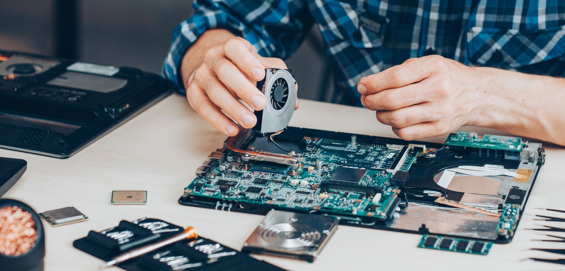 Technician repairs an open laptop, holding a cooling fan above the motherboard