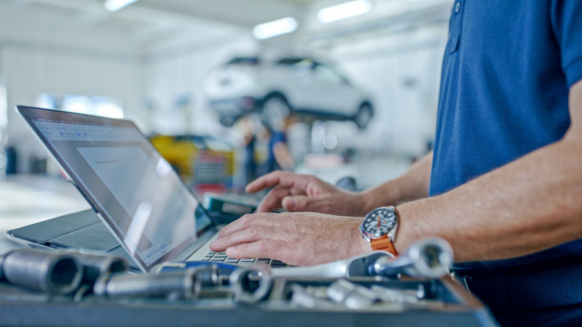 Mechanic using laptop in auto repair shop with car on lift in background and tools on table.
