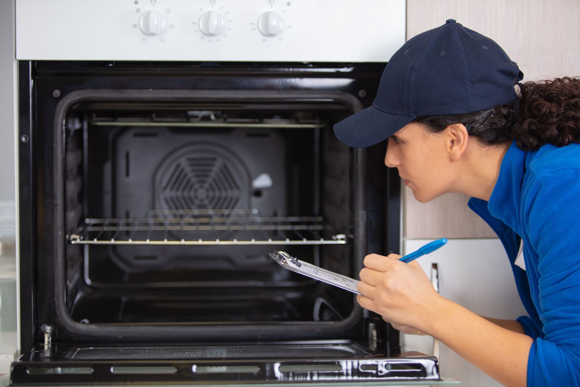 A woman is looking into an oven and writing on a clipboard.
