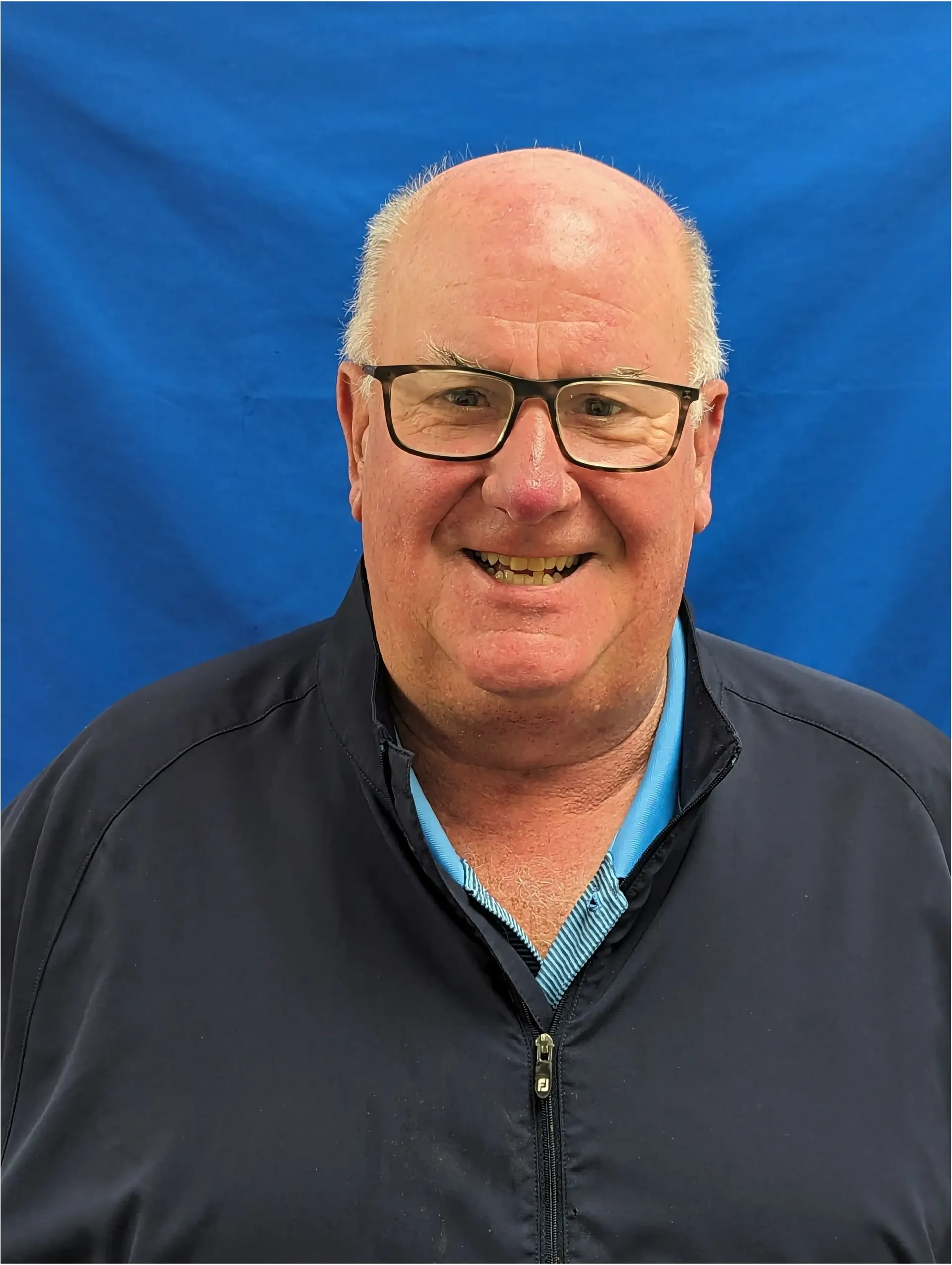 A bald man wearing glasses and a blue shirt is smiling in front of a blue background.