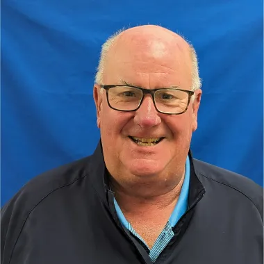 A bald man wearing glasses and a blue shirt is smiling in front of a blue background.