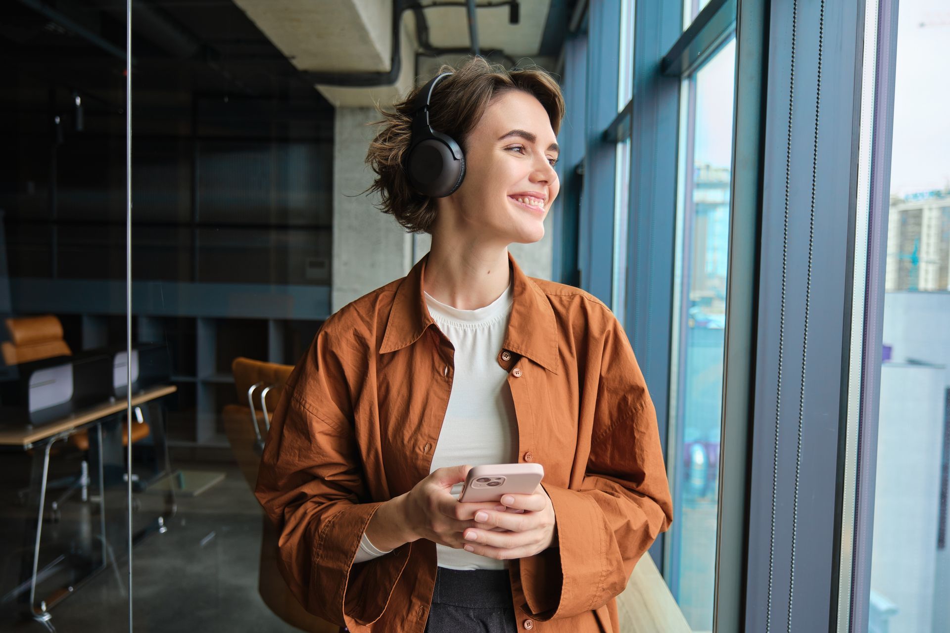 A woman wearing headphones is looking out of a window while holding a cell phone.