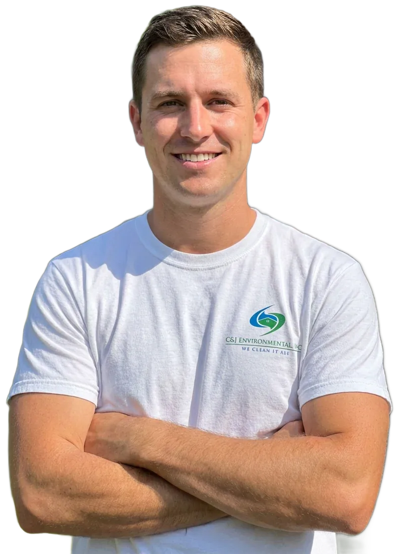 Man with arms crossed, wearing safety glasses and apron, in a wood shop, smiling.