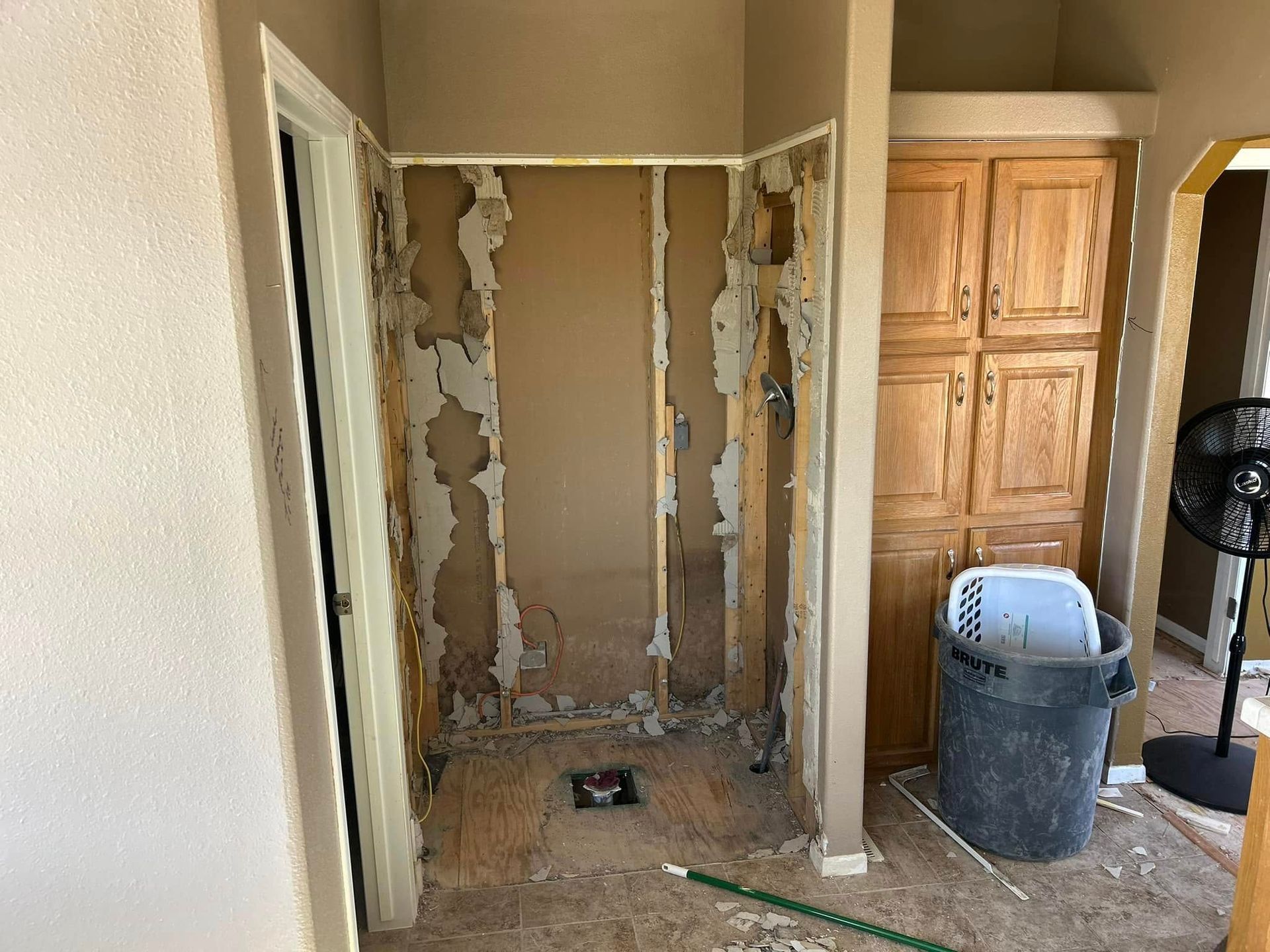 Bathroom demolition: walls stripped to studs, debris on floor, door frame on left, cabinet on right, trash can present.