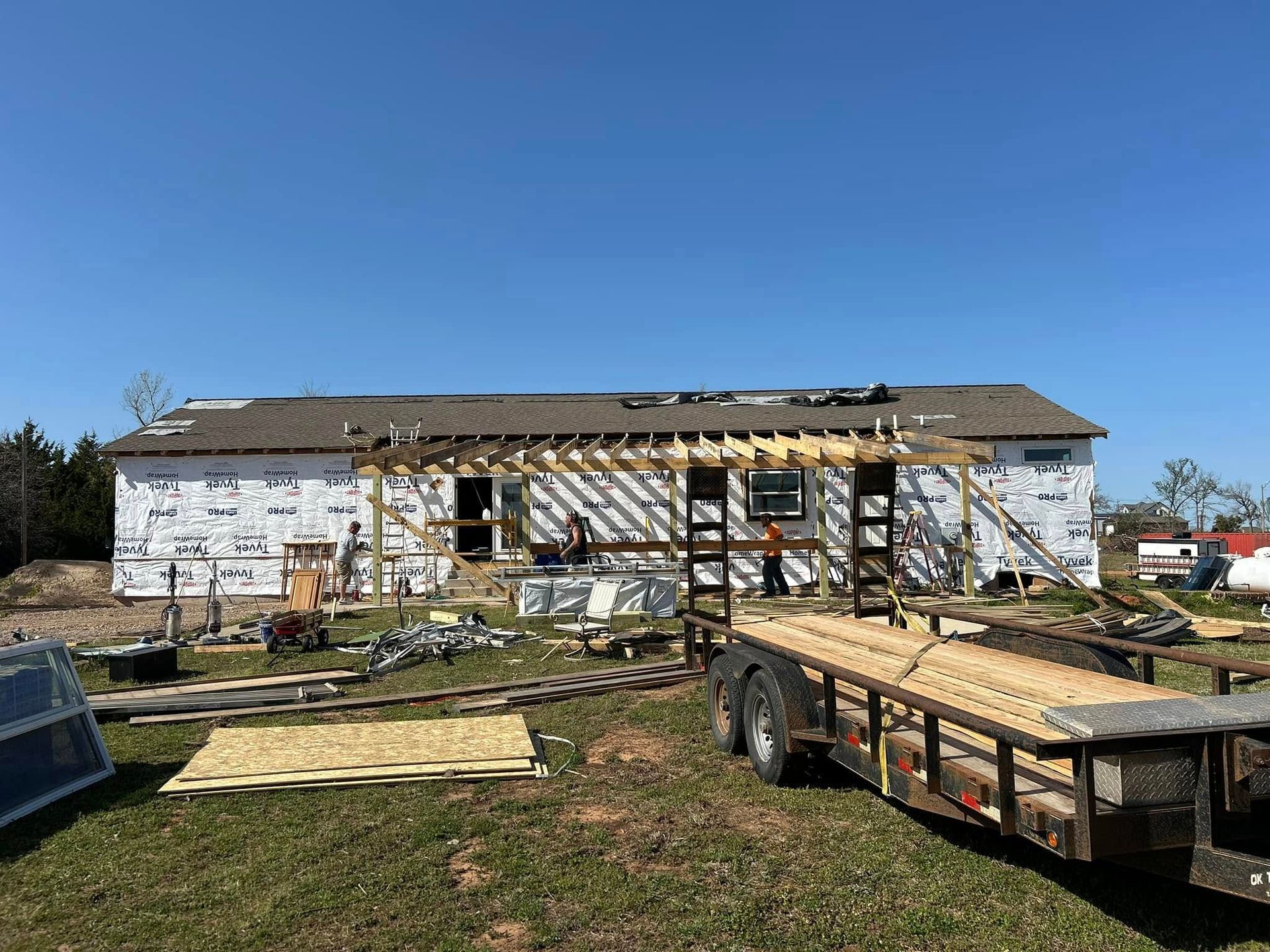 Construction site of a house under renovation, white siding, brown roof, trailer in the foreground, clear blue sky.