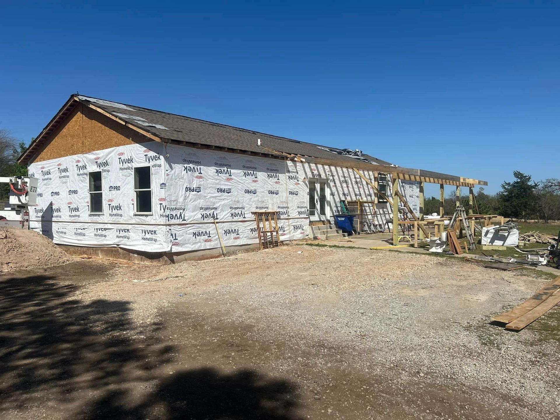 House under construction, exterior view. Building wrapped in white material, gravel ground, clear blue sky.