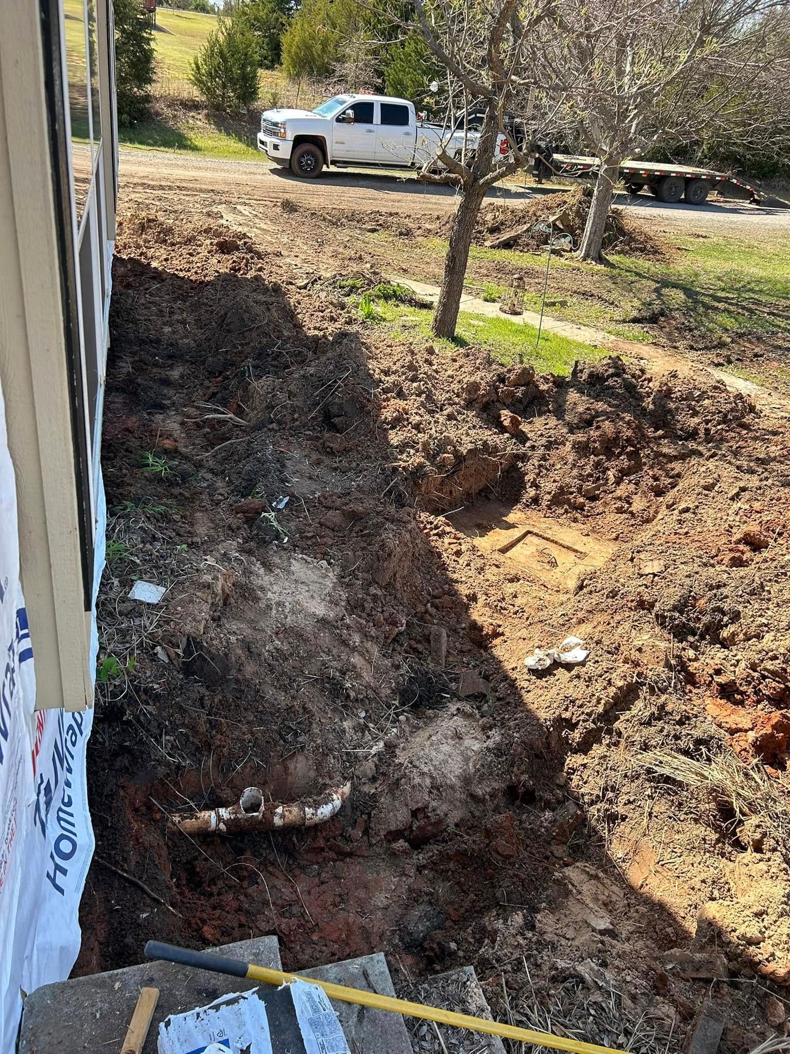 A trench dug along a house foundation, exposing plumbing. A white truck is visible in the background.