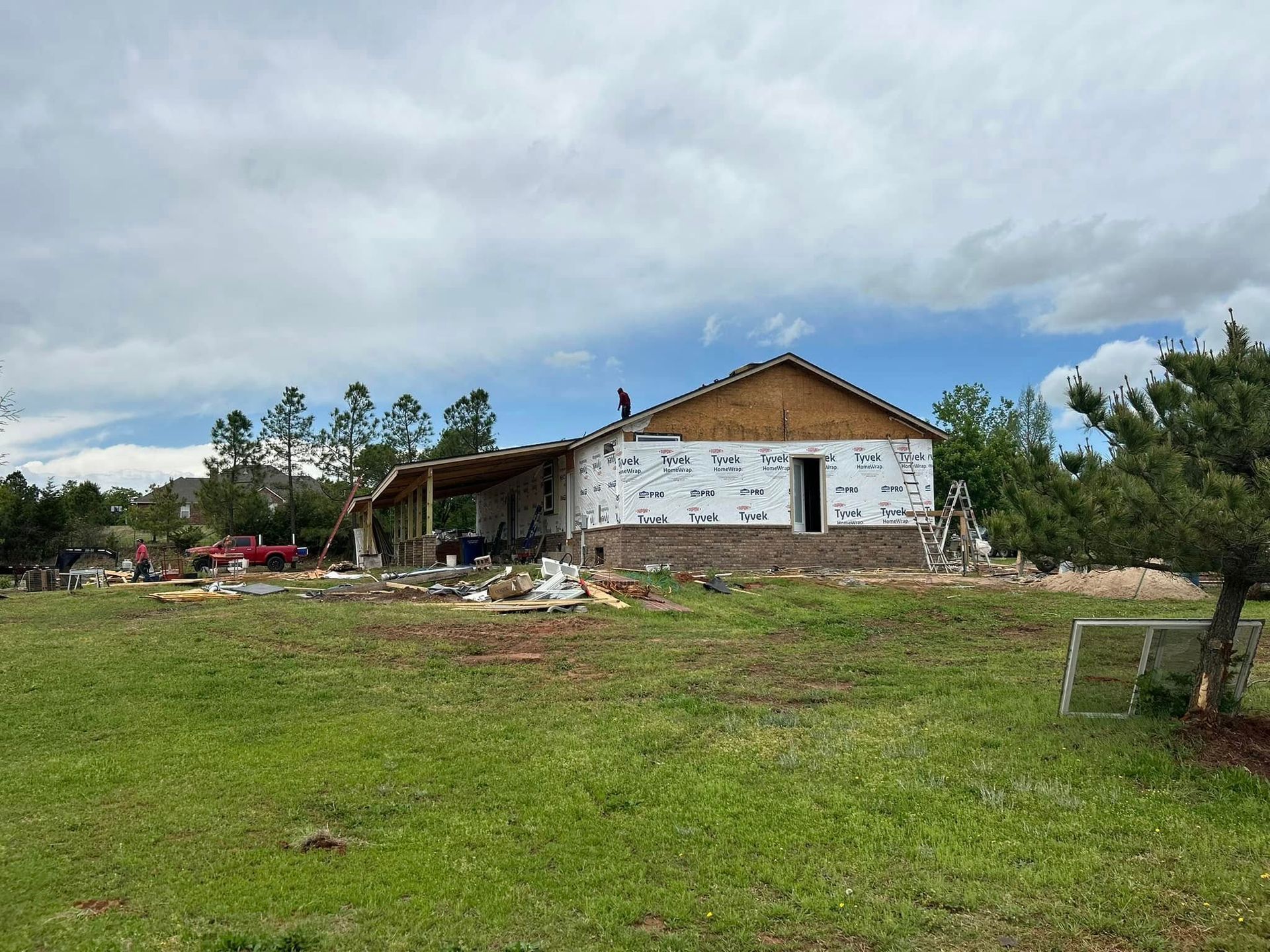 House under construction, partially covered in white wrap, set in a grassy field under cloudy skies.