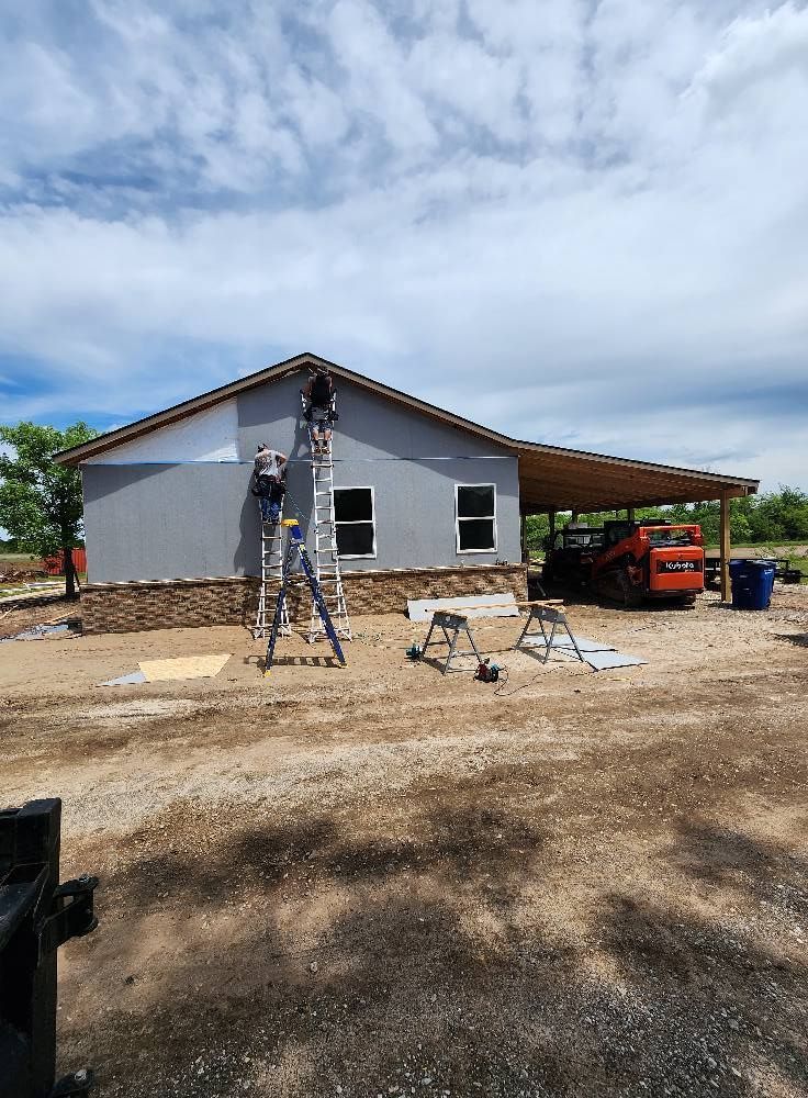 Construction workers on ladders installing siding on a gray house under a blue sky with a skid steer in the background.