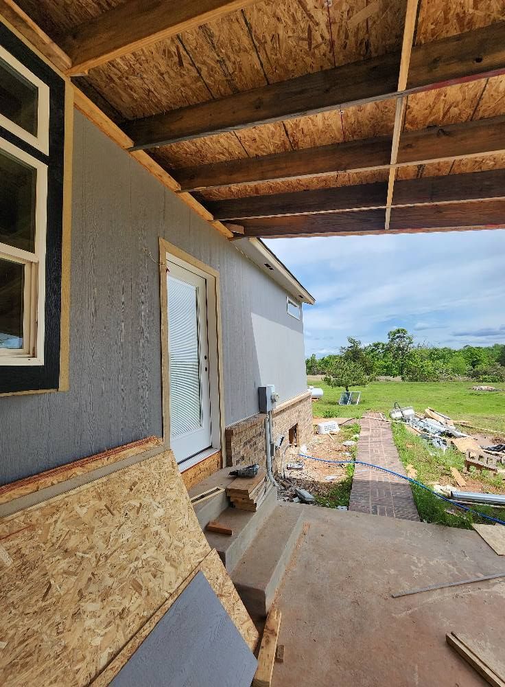 Construction site with front door, gray siding, and exposed wooden beams. Overcast sky, rural setting.
