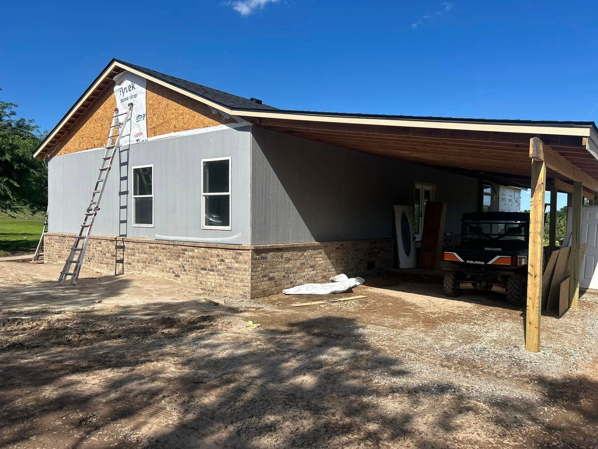 A partially built house with gray siding, stone base, and a carport; a ladder leans against the side.