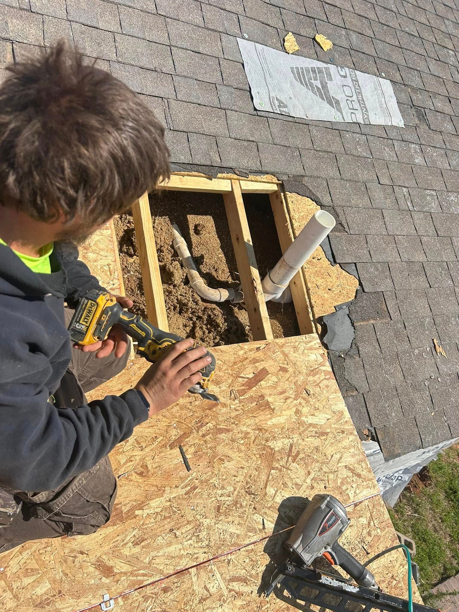 Roofer using a nail gun to repair a roof with exposed framing and a white pipe.