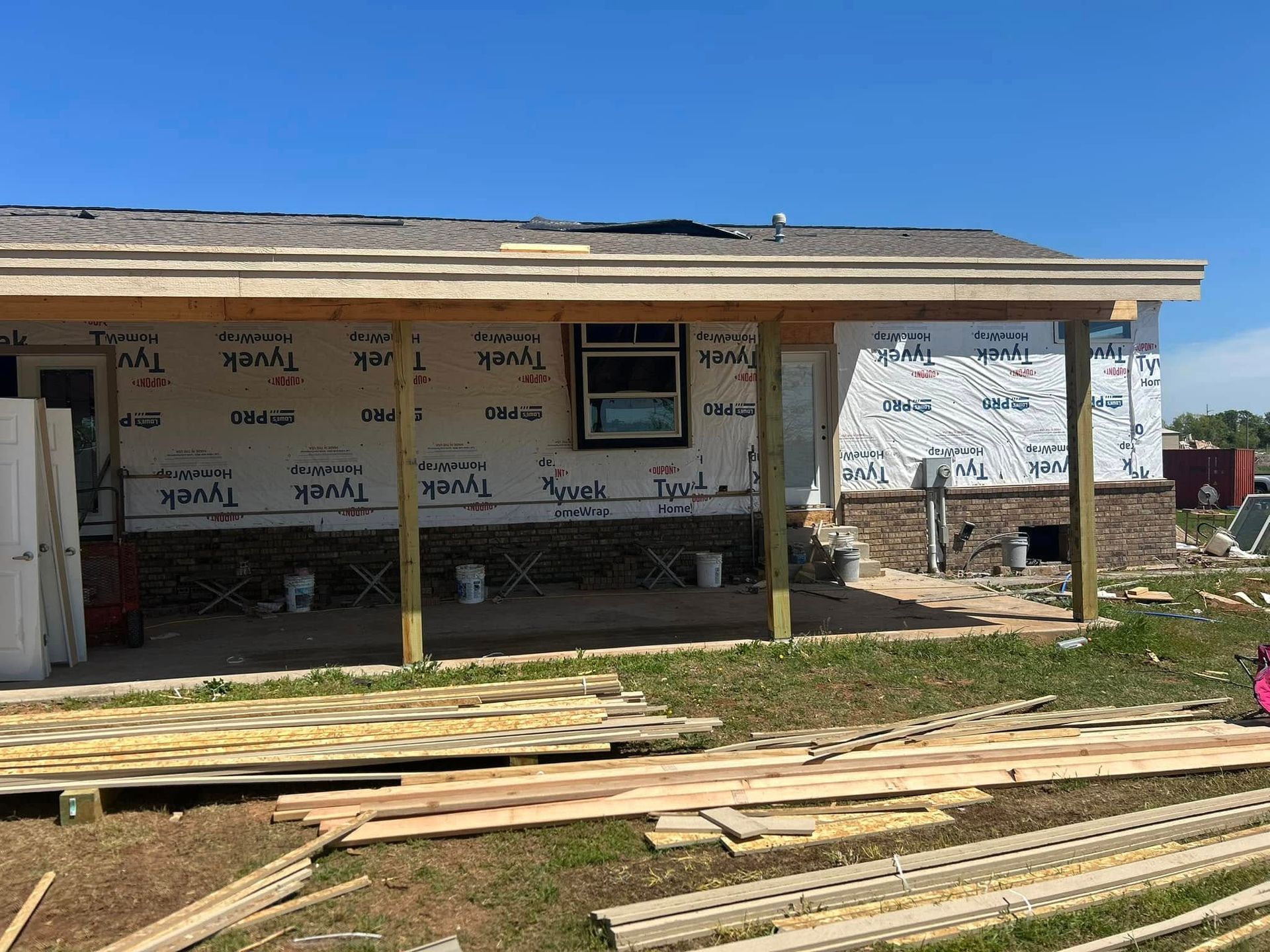 Construction site: House with wooden porch, covered in Tyvek, and surrounded by building materials. Blue sky.
