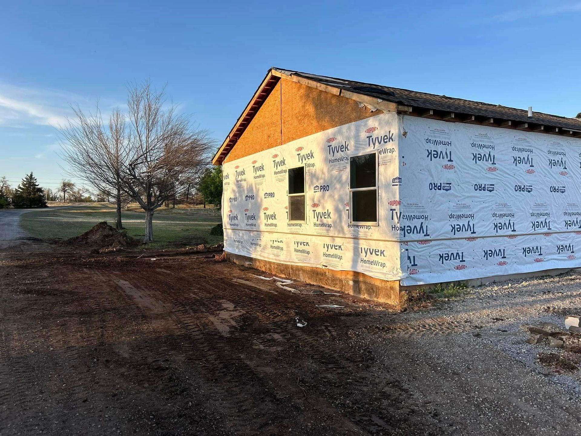 A partially constructed building with Tyvek wrap, two windows, and a dirt lot in front.