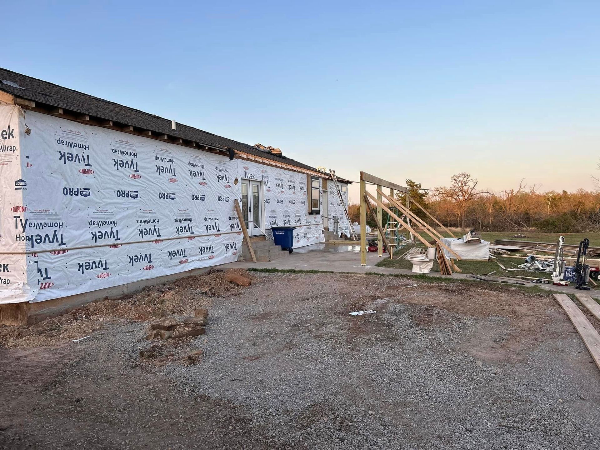 Construction site: building with Tyvek wrap, wooden stairs, gravel yard, and blue sky.