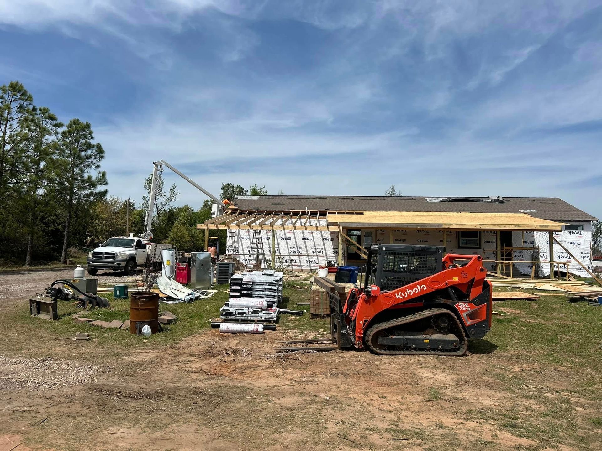 Construction site with a skid steer, truck, and materials; house partially framed.