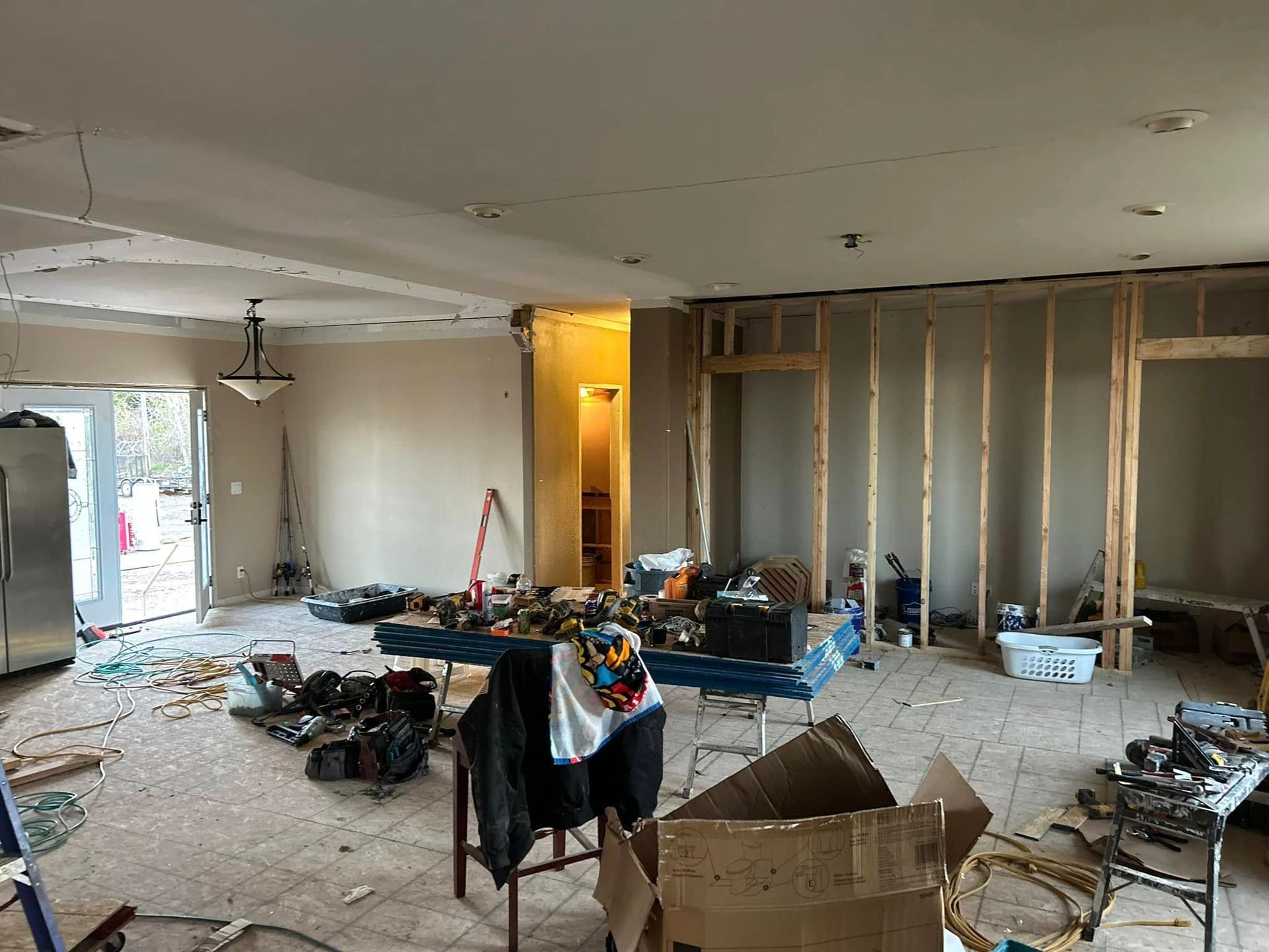 Living room under renovation: exposed framing, tools and debris scattered, doorway to outdoors on left.