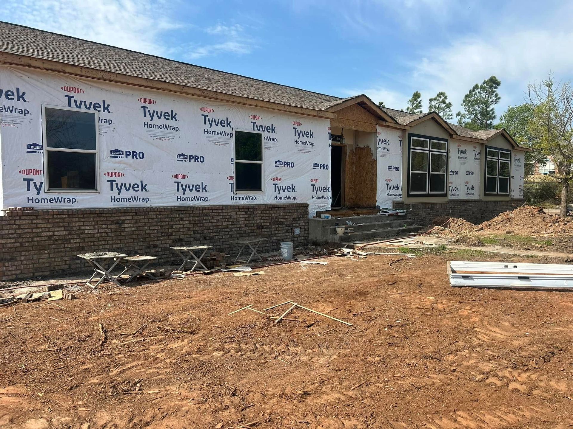 House under construction, with Tyvek wrap, brickwork, and a dirt lot on a sunny day.