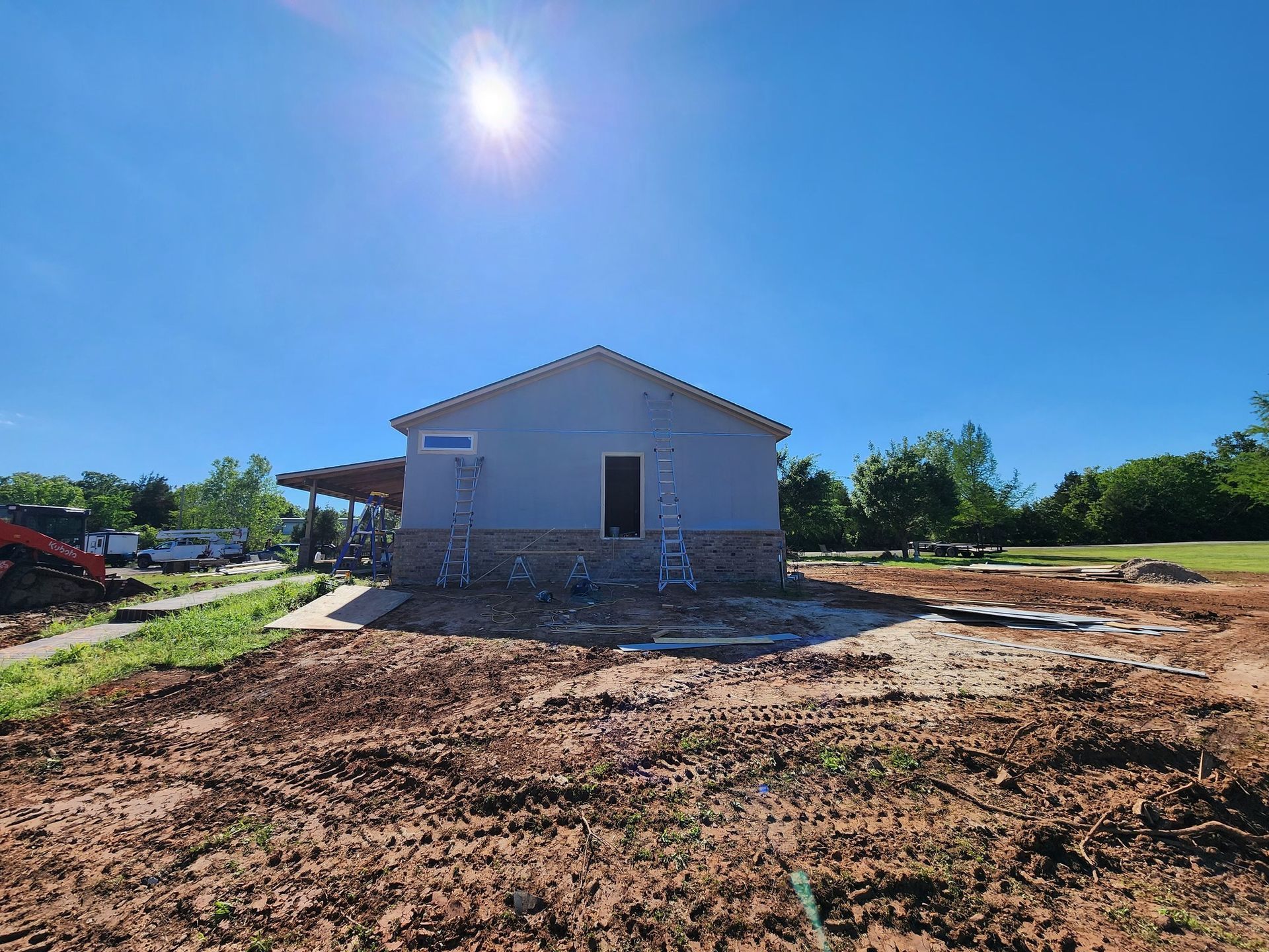 Building under construction, gray siding, stone base, blue sky, sunny day, and dirt lot.