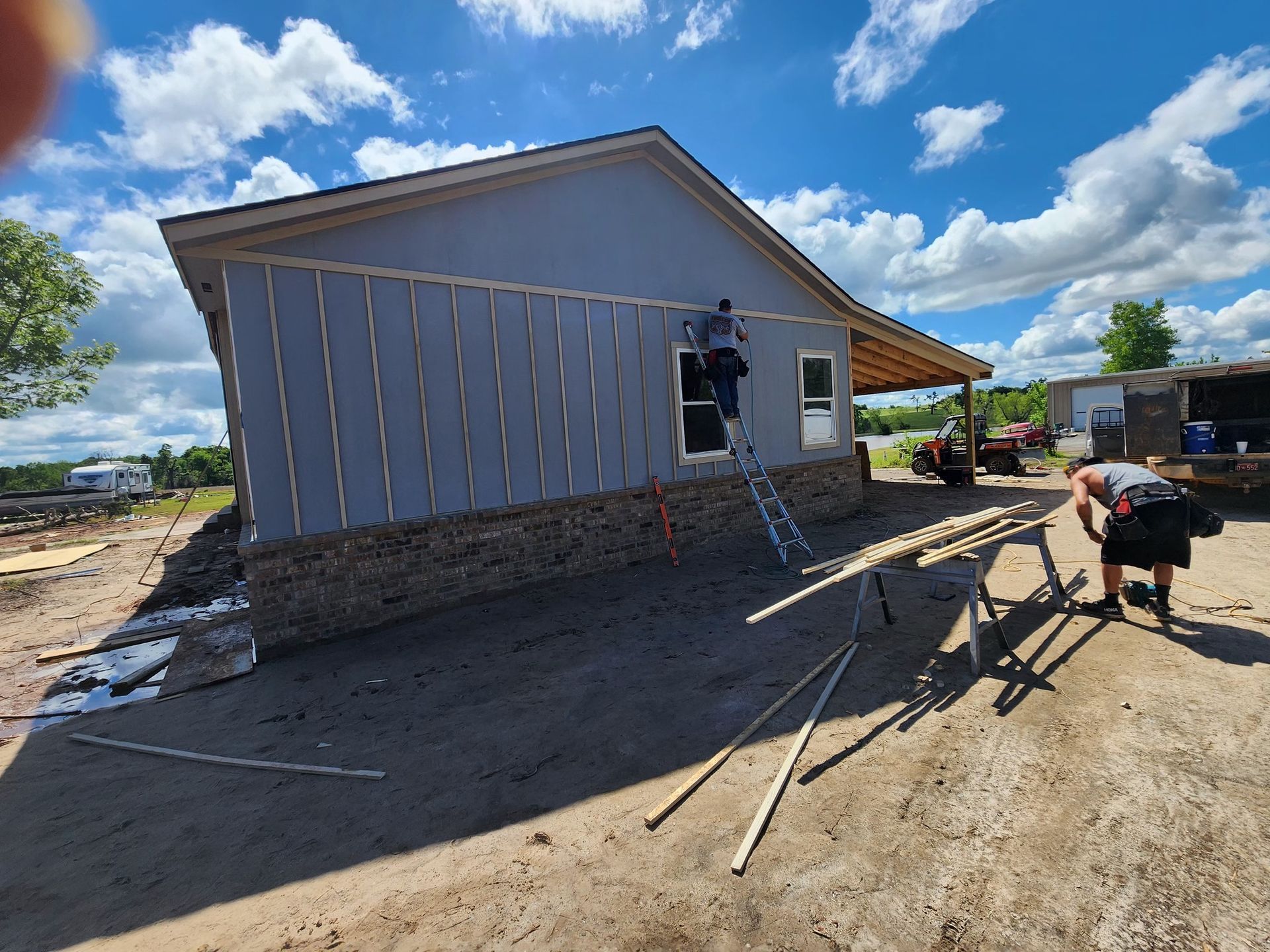 Construction workers siding a house with blue siding and brick base on a sunny day.
