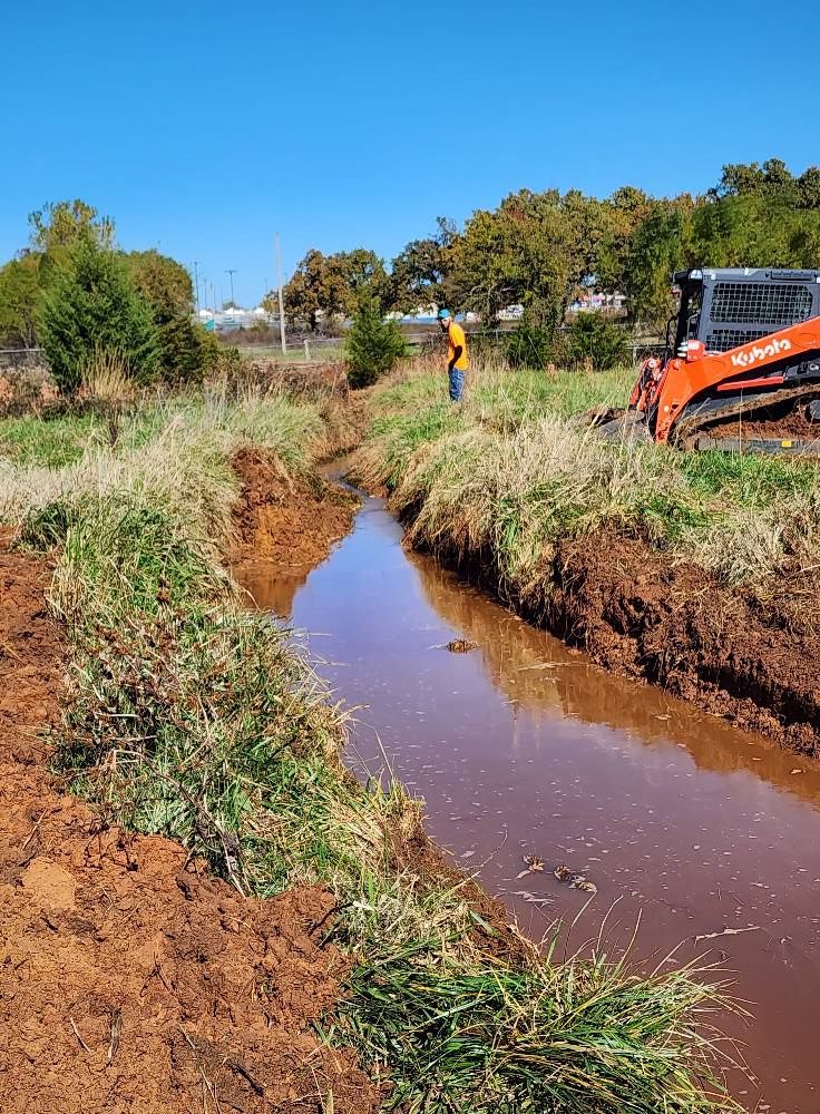 A small muddy creek with a person and a construction vehicle working on the banks.