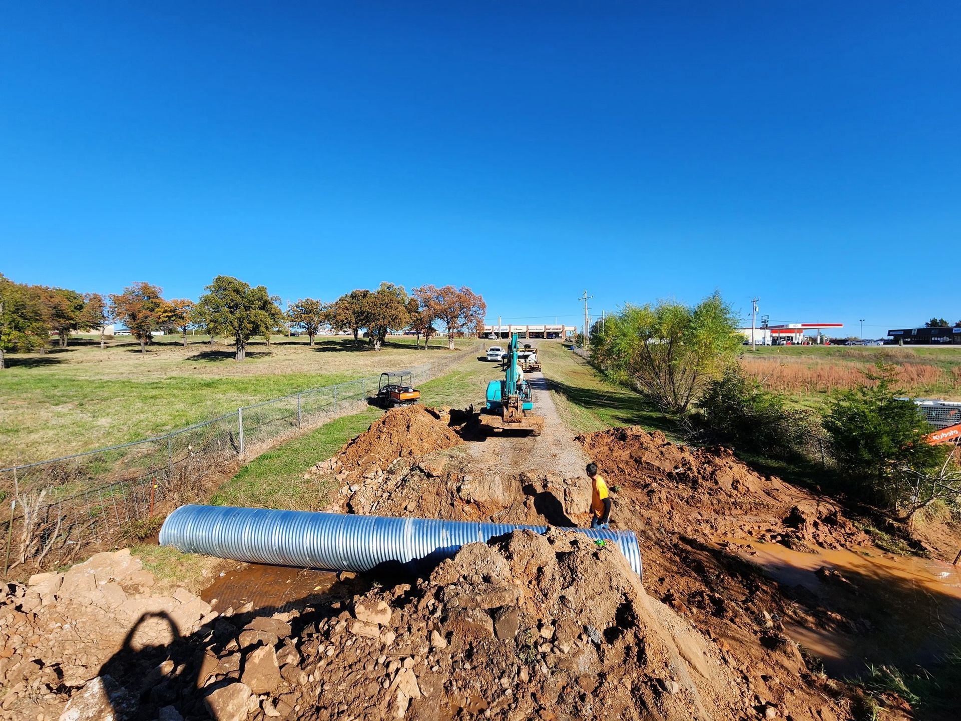 Construction site with a large metal pipe being installed in a trench along a grassy area under a blue sky.