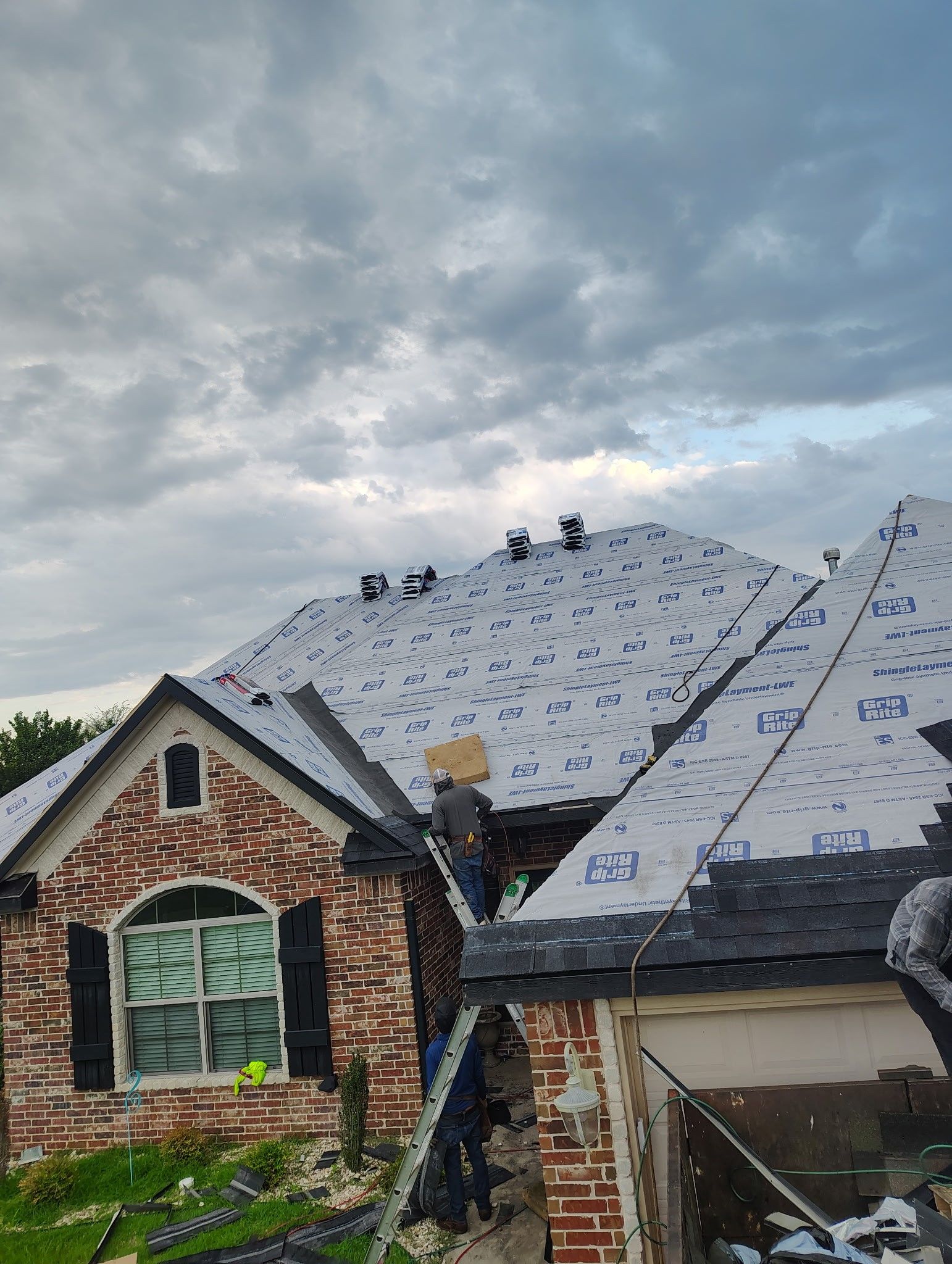 Workers installing shingles on a brick house roof, cloudy sky overhead.