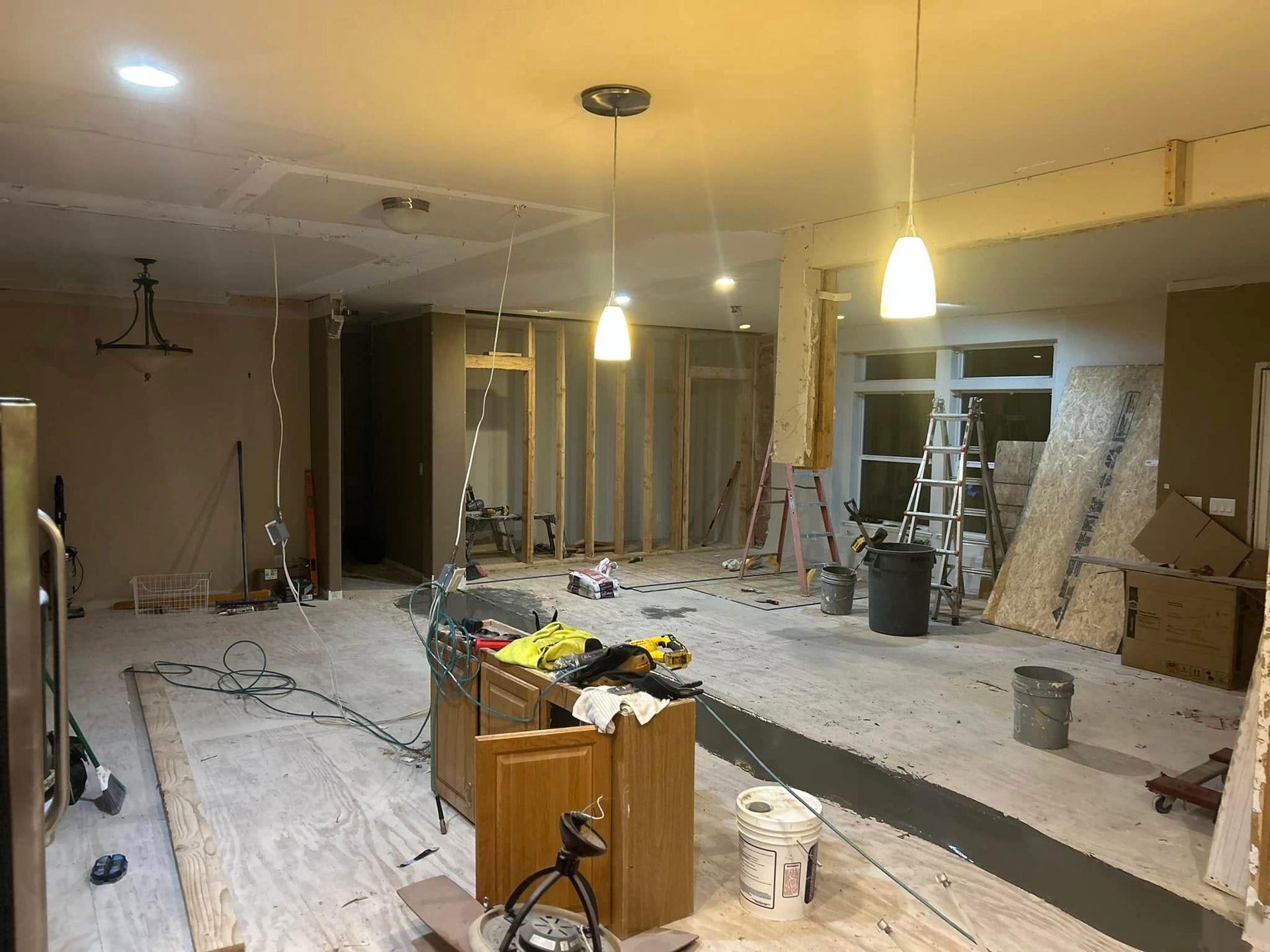 Interior renovation: walls opened, kitchen cabinets in foreground, ladders, tools, and debris scattered.